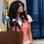 young brown skinned woman in pink sweater vest stands at podium in front of flags presenting