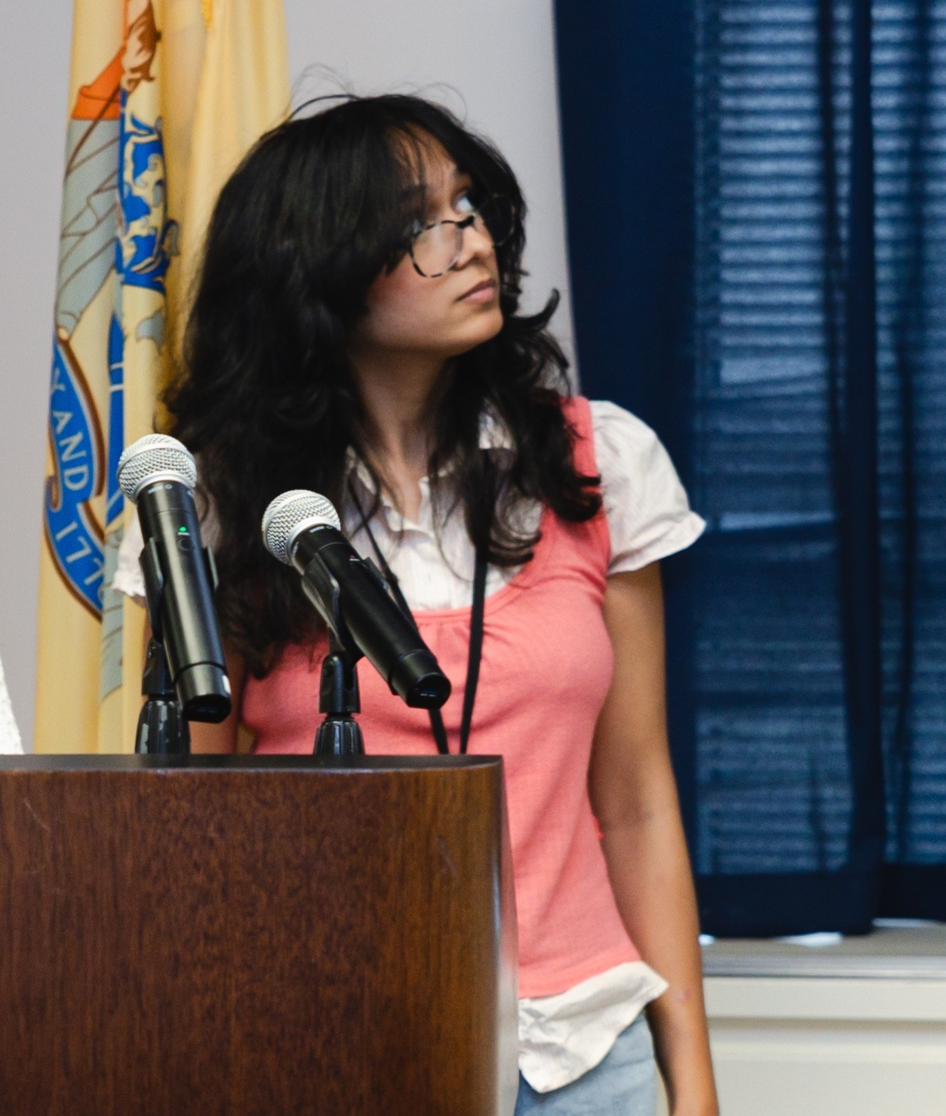 young brown skinned woman in pink sweater vest stands at podium in front of flags presenting